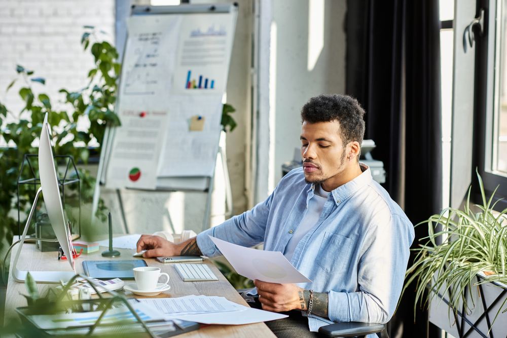 A man sits at a desk in a bright office, looking at a piece of paper. A computer, coffee cup, and documents are on the desk. A flip chart with graphs is in the background, surrounded by green plants.