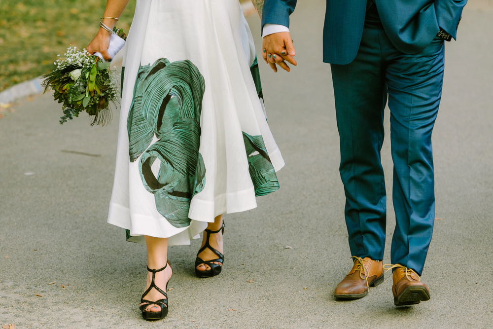 A couple walk hand in hand; the woman wears a white dress with green patterns and black heels, holding a bouquet, whilst the man wears a blue suit and brown shoes.