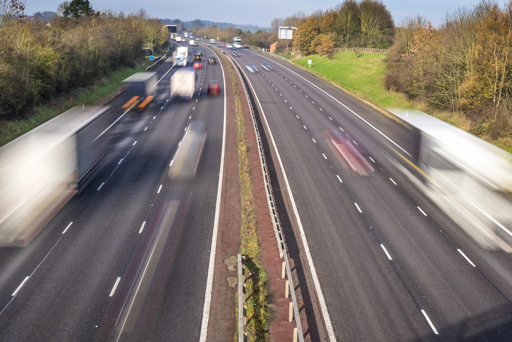 A motorway with several cars and lorries travelling fast in both directions. The vehicles appear blurred due to motion, and there are trees lining both sides of the road.
