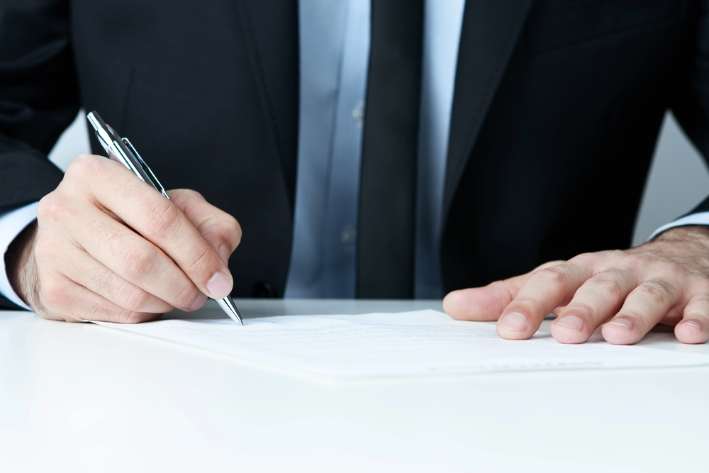 A person in a suit sits at a desk, holding a pen and signing a document with one hand whilst the other hand rests on the paper.