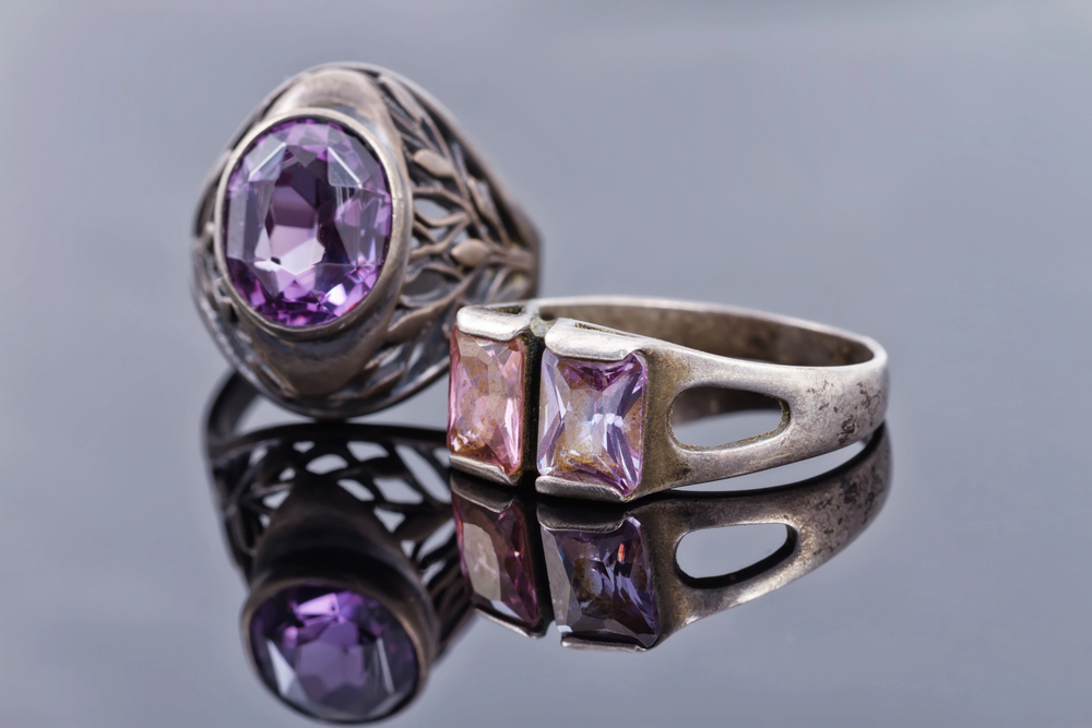 Two silver rings with purple gemstones rest on a reflective surface. One ring features a large oval stone in a detailed setting, whilst the other has two square-cut stones with an open band design.