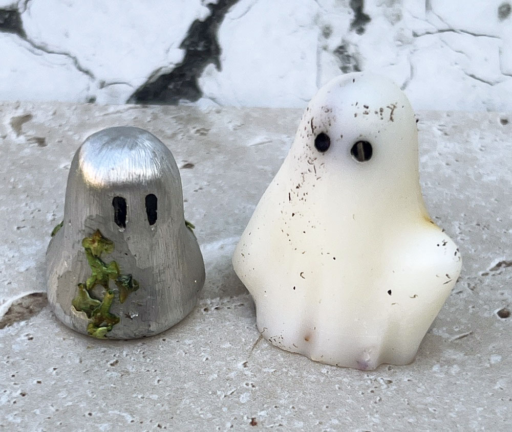 Two small ghost figurines, one metallic with green residue and one white with black spots, sit side by side on a stone surface in front of a marbled background.