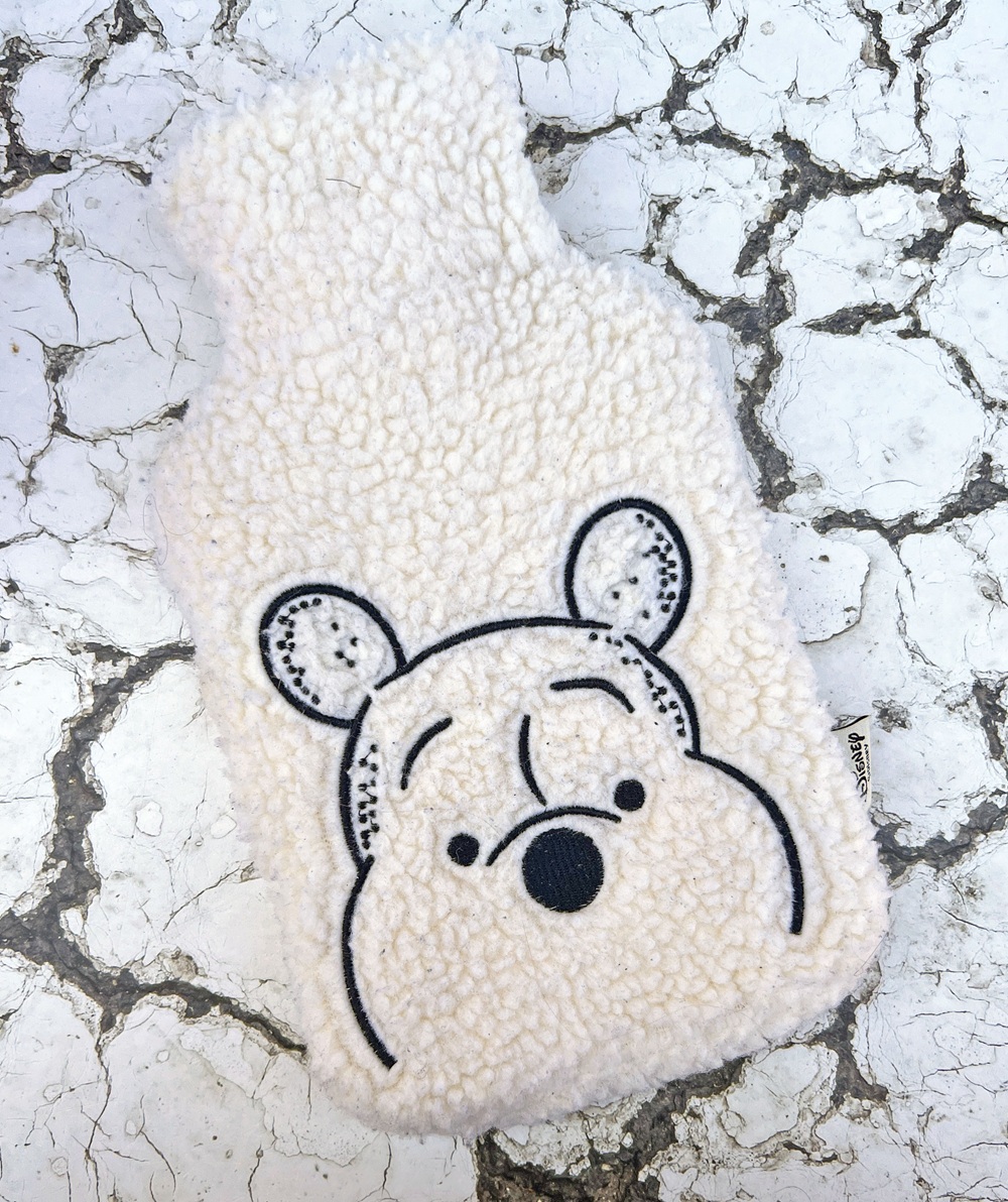 A fuzzy hot water bottle cover with an embroidered bear face, placed on cracked white stone pavement.