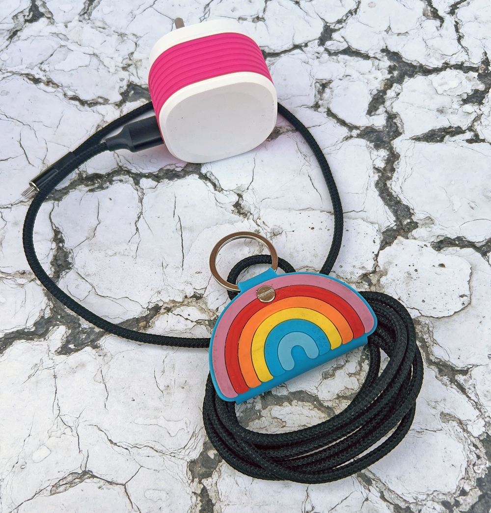 A black charging cable is coiled and held with a rainbow-shaped leather holder. A pink and white plug is attached to the cable, all placed on a cracked white surface.