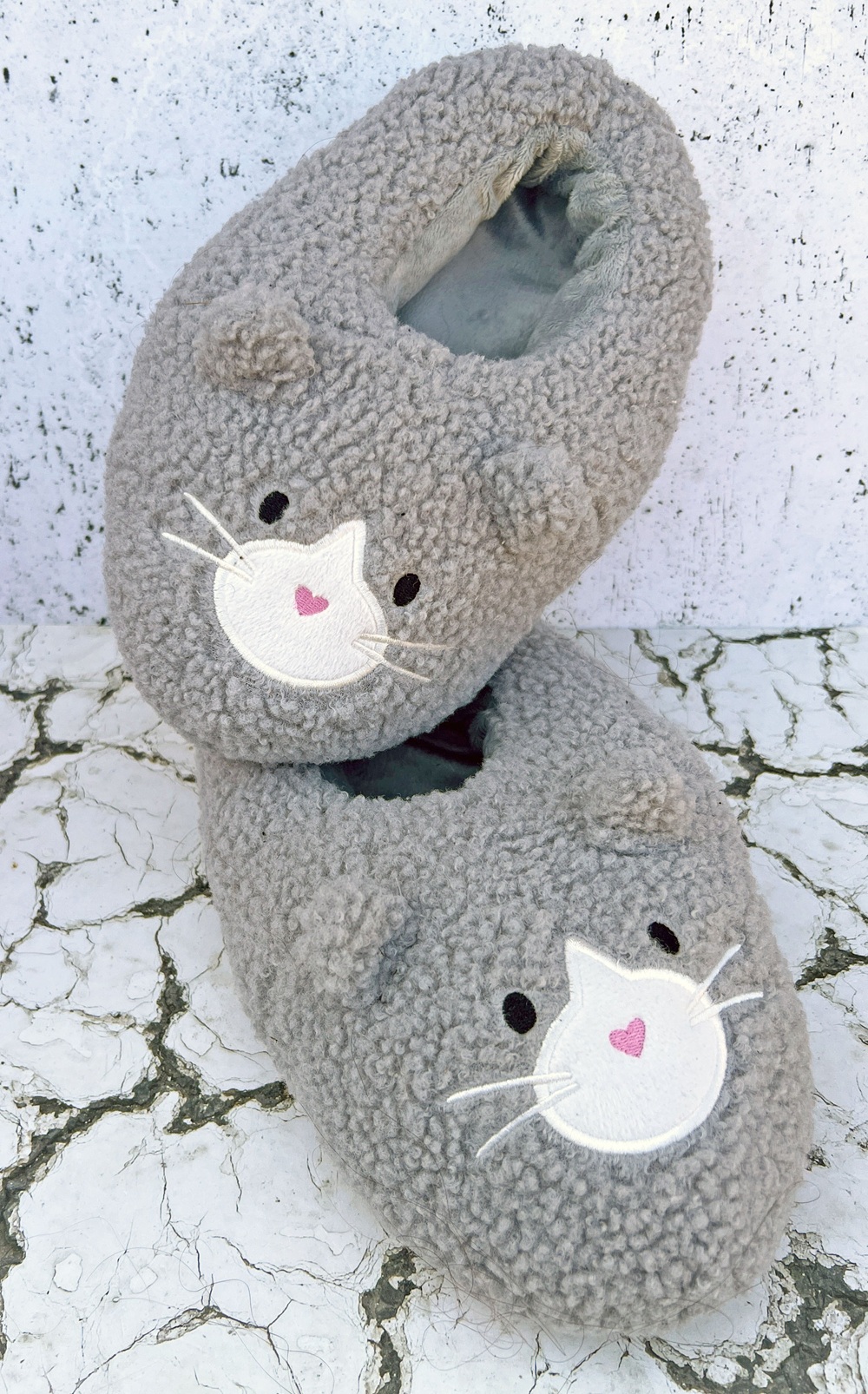 Two fuzzy grey slippers with embroidered cat faces featuring white whiskers, pink heart-shaped noses, and small ears. They are stacked on a cracked white surface against a textured white background.