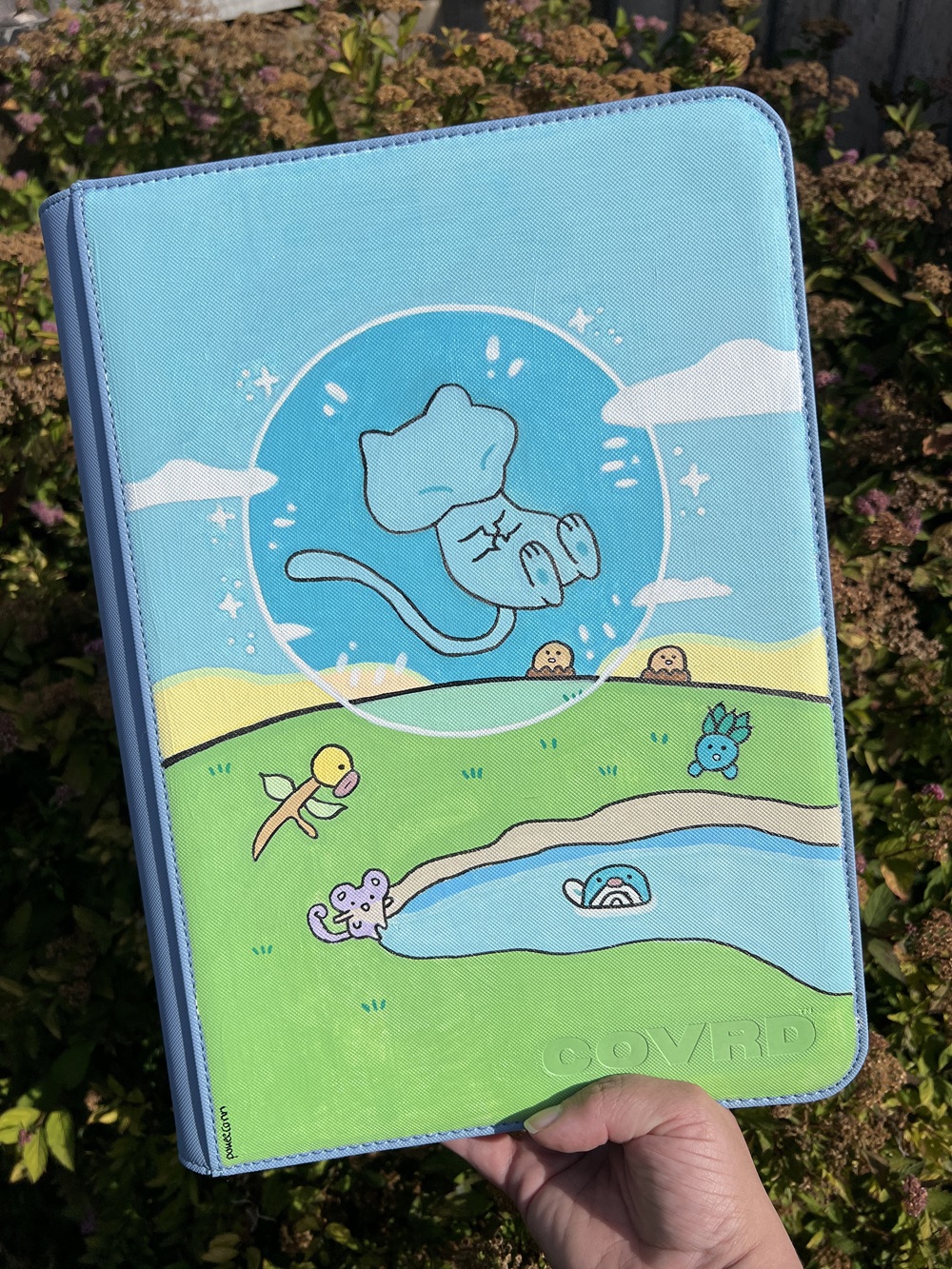 A hand holds a blue case with cartoon-style art of a floating blue cat, cute animals, a pond, and grassy hills under a bright blue sky with clouds.