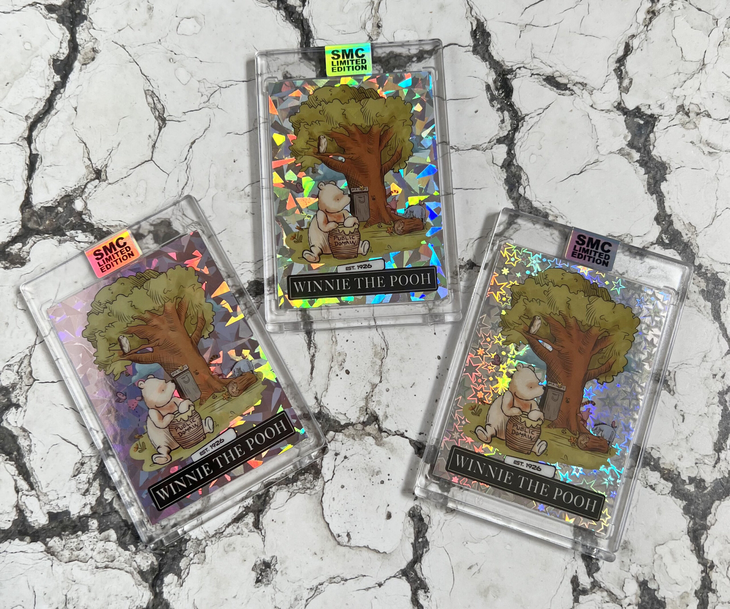 Three limited edition holographic Winnie-the-Pooh cards in protective cases are displayed on a marbled surface. Each card shows Pooh sitting under a tree, with reflective rainbow patterns and SMC Limited Edition labels.