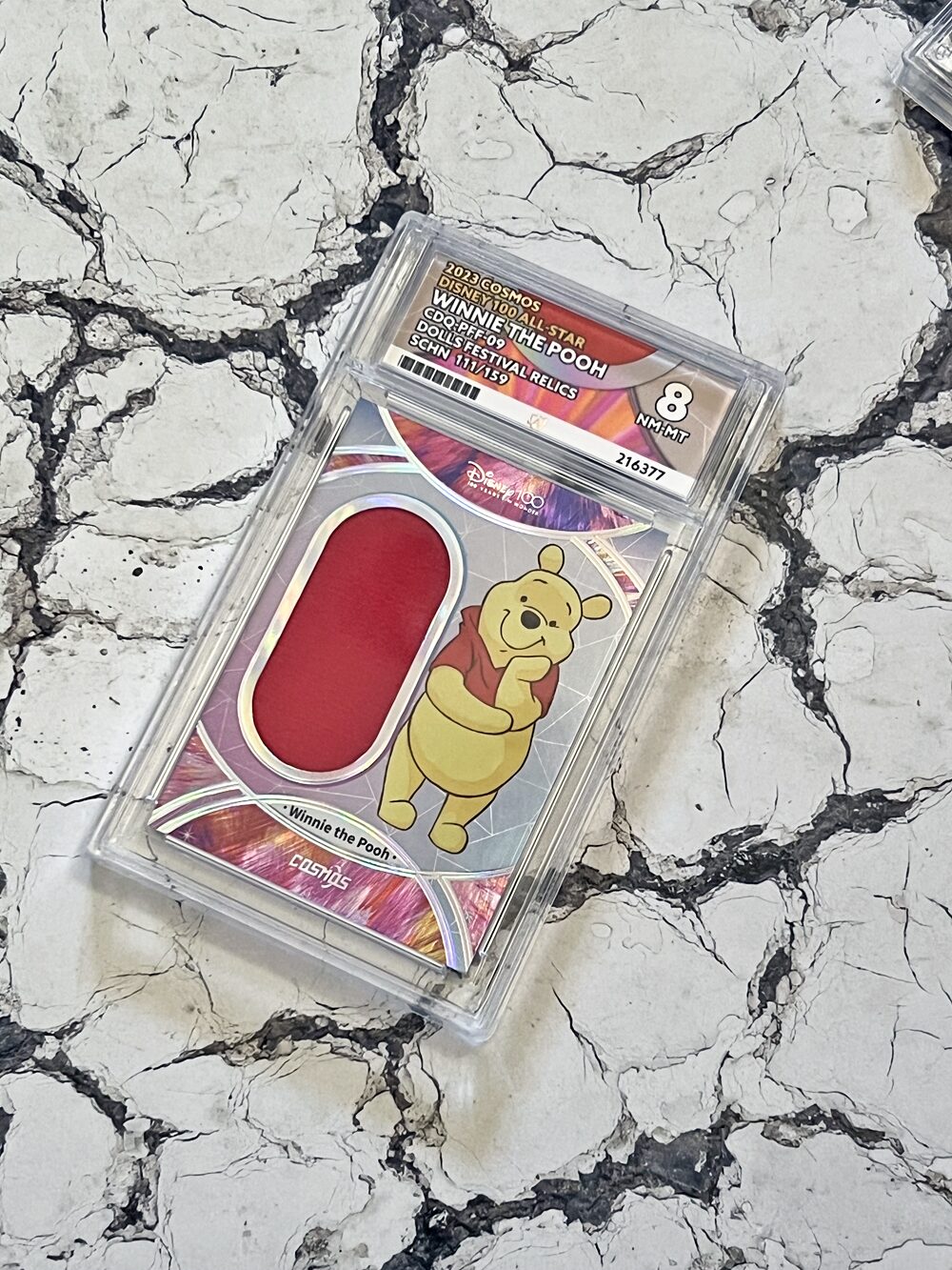 A graded Disney Winnie the Pooh trading card with a red patch is encased in plastic and placed on a cracked marble surface.