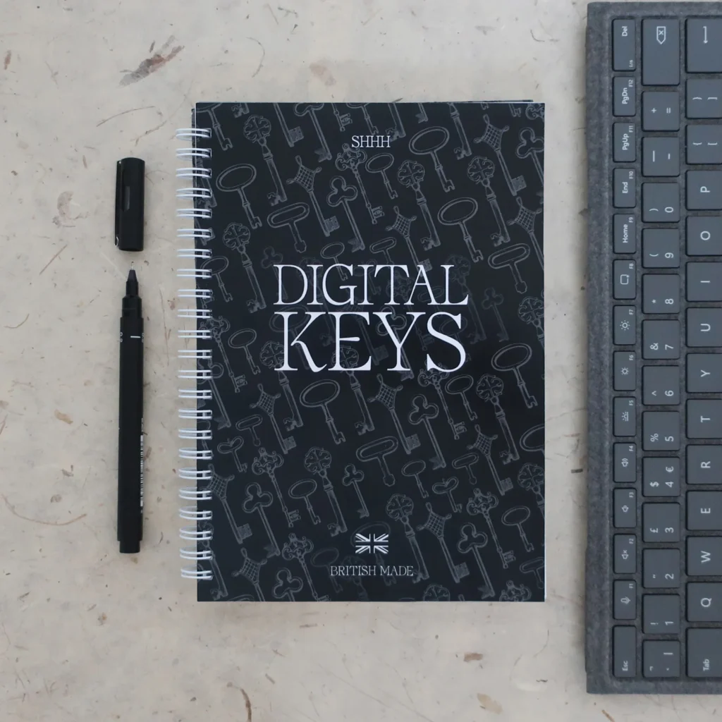 A spiral notebook labelled DIGITAL KEYS sits on a desk next to a black pen with its cap off and the edge of a computer keyboard. The notebook cover features various key illustrations and text BRITISH MADE.