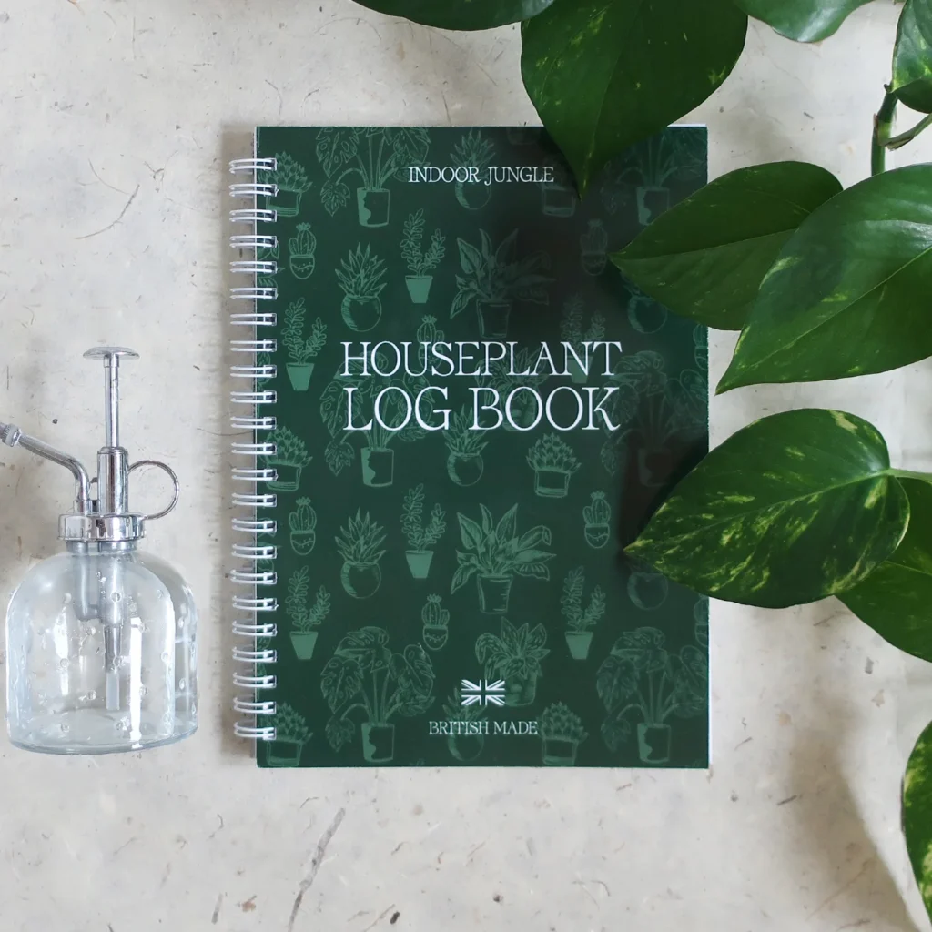 A green Houseplant Log Book with plant illustrations lies on a light surface, next to a glass spray bottle and green plant leaves.