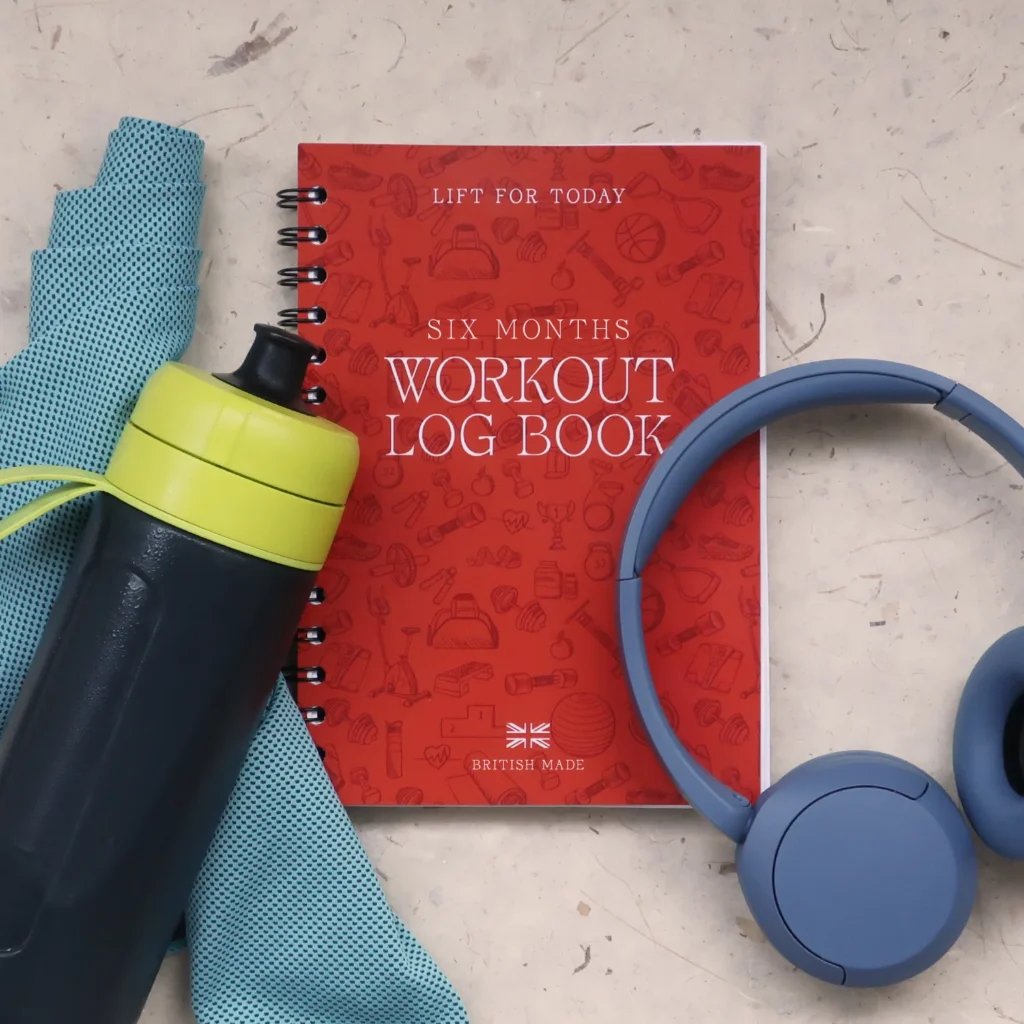 A red Six Months Workout Log Book sits on a light surface with a blue towel, yellow-lid water bottle, and blue headphones next to it.