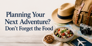 A wooden table with a bowl of salad, almonds, a map, camera, hat, rucksack, and toy aeroplane beside text: Planning Your Next Adventure? Don’t Forget the Food.
