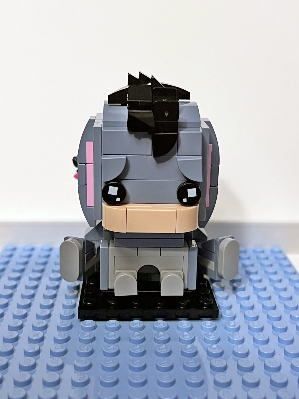 A Lego-style brick figure resembling Eeyore, the donkey from Winnie the Pooh, sits on a blue baseplate. The figure is mostly grey with black hair, pink cheeks, and droopy ears.