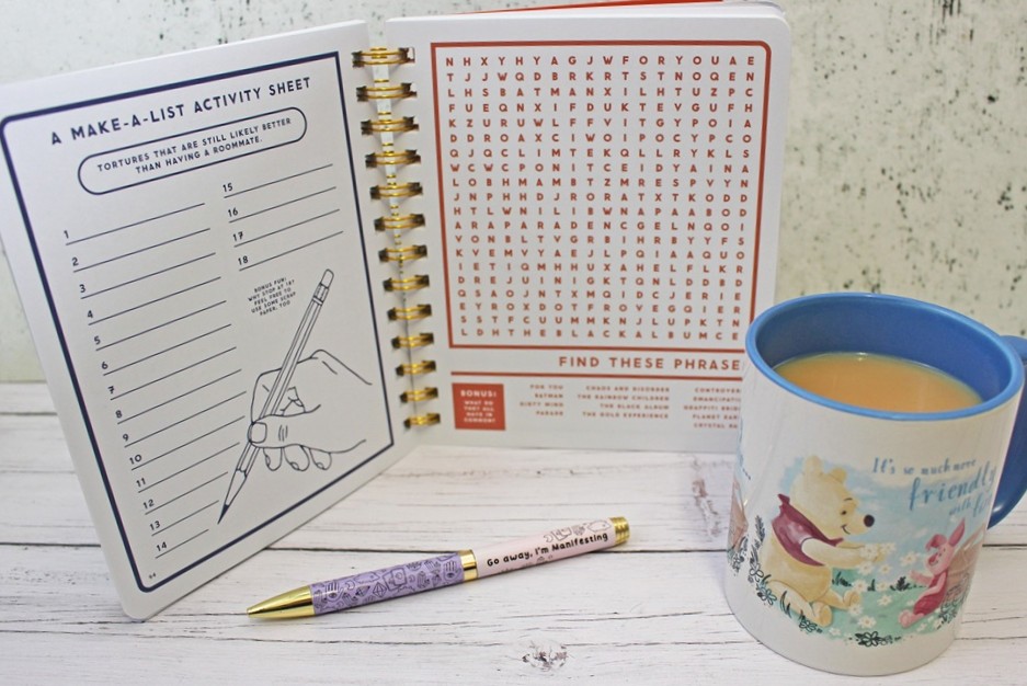 Open exercise book with activity sheet and wordsearch, two pens, and a mug of tea with a bear illustration and text: It’s so much friendlier… on a white wooden surface.