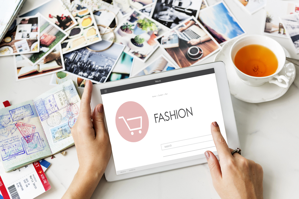 A person uses a tablet displaying a fashion shopping site. Nearby are a cup of tea, a notebook, and scattered photos on a white table.