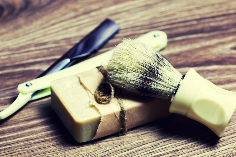A shaving brush and a bar of soap rest on a wooden surface, with a cut-throat razor placed in the background.