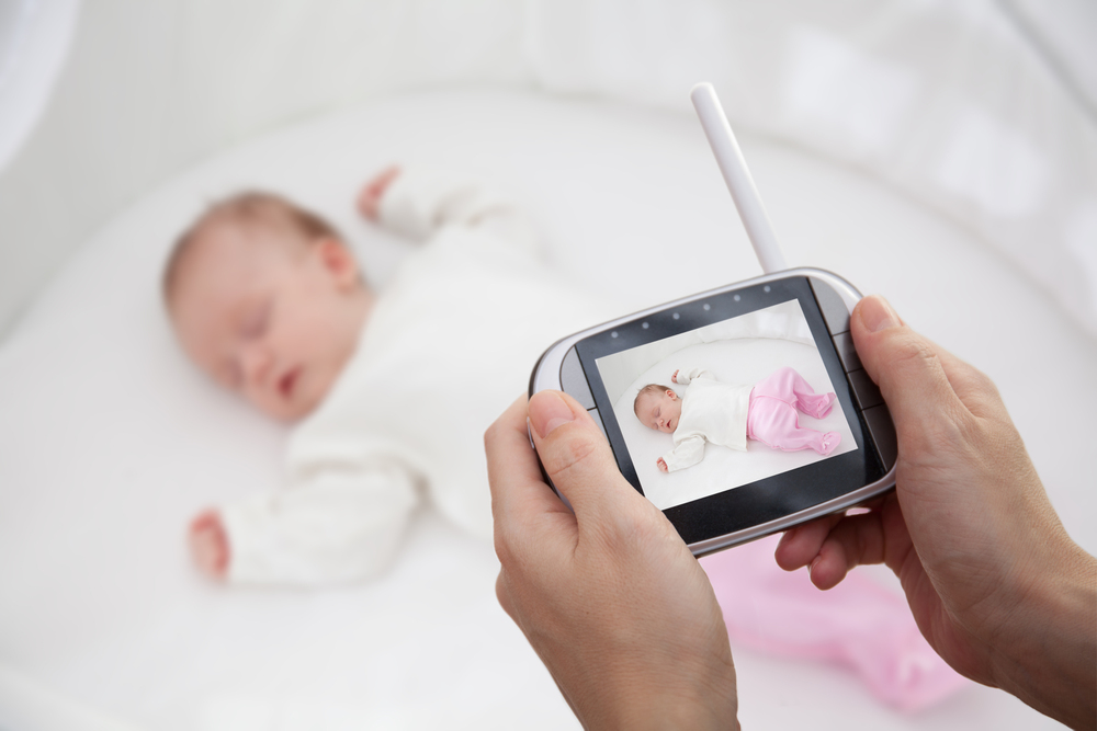 A person holds a baby monitor displaying a video of a sleeping infant dressed in white and pink, lying on their back in a white cot.