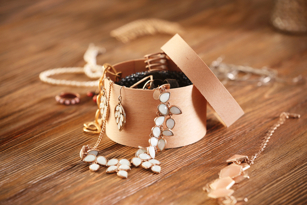 A round, light brown box with the lid off, filled with assorted jewellery including a beaded bracelet, a leaf earring, and a floral necklace, on a wooden surface.