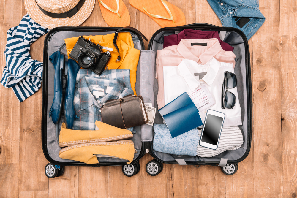 Open suitcase on a wooden floor, packed with clothes, shoes, camera, sunglasses, passport, boarding pass, and a mobile phone, with a hat, flip-flops, and a striped shirt nearby.
