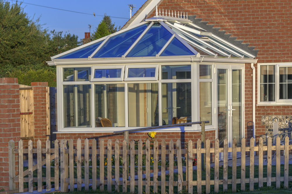A glass conservatory with a blue-tinted roof is attached to a brick house. There is a wooden fence and garden with a yellow flower in the foreground.