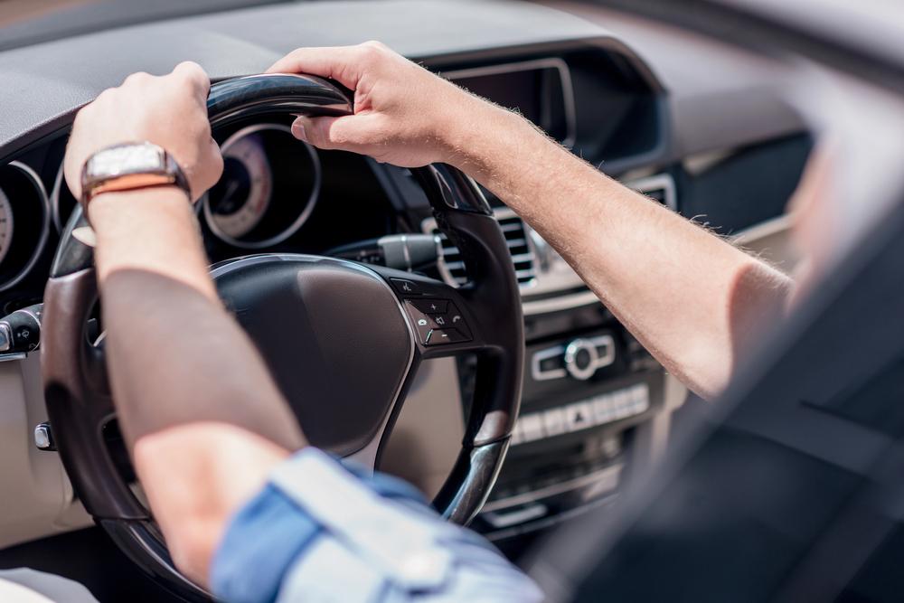 A person with a watch on their left wrist is holding a black steering wheel and driving a car, with the dashboard and control buttons visible.