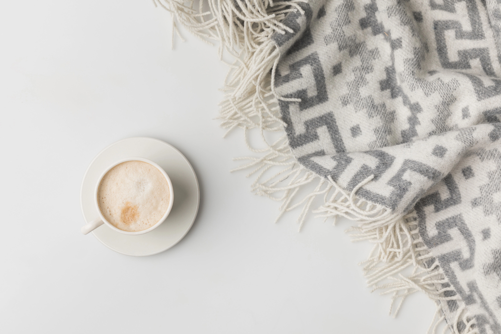 A cup of coffee with frothy milk sits on a saucer next to a soft, grey-and-white patterned blanket with fringe, on a white surface.