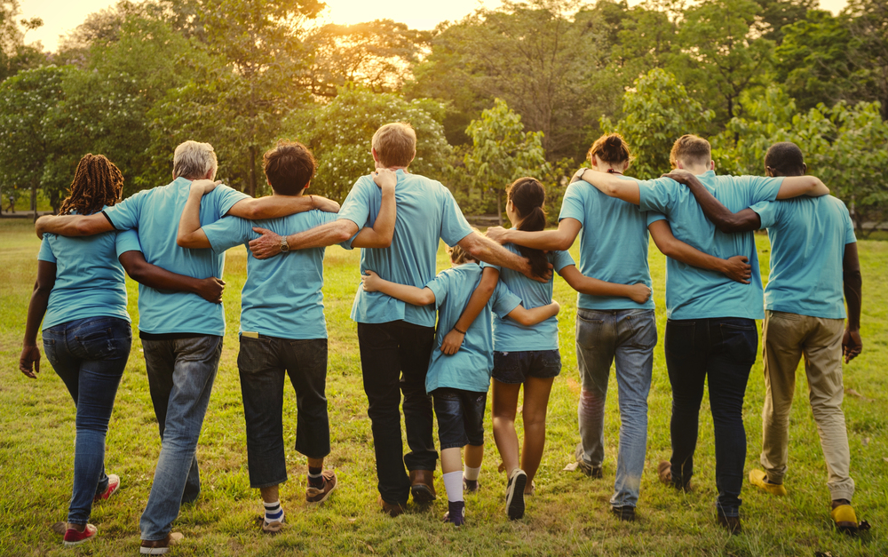 A diverse group of people in blue shirts stand outside with arms around each other's shoulders, facing away from the camera, in a grassy park with trees and sunlight in the background.