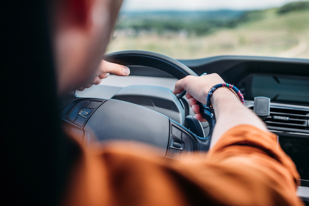 A person in an orange shirt is driving a car, gripping the steering wheel with both hands. The photo is taken from behind, showing part of the dashboard and a blurred outdoor landscape through the windscreen.