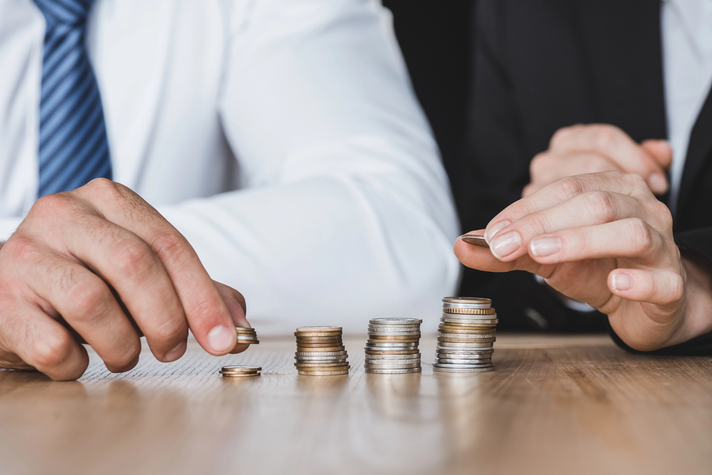 Two people in business attire stack and organise coins on a wooden table, arranging several neat piles of coins of varying heights.