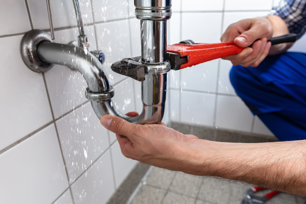 A person uses a spanner to mend a leaking pipe under a sink, with water spraying from the joint. The scene takes place against a tiled wall in a bathroom or kitchen.