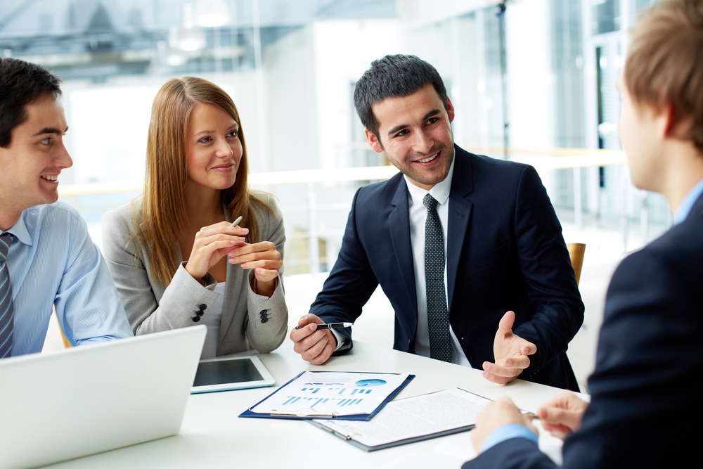 Four professionals in business attire sit at a table with documents and a laptop, engaged in a discussion and smiling in a bright office setting.