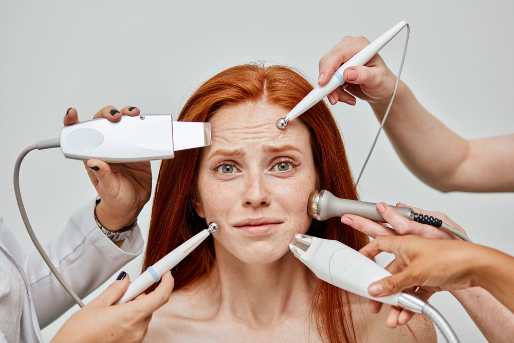 A red-haired woman looks uneasy as multiple hands hold various skincare and beauty devices to her face against a plain background.