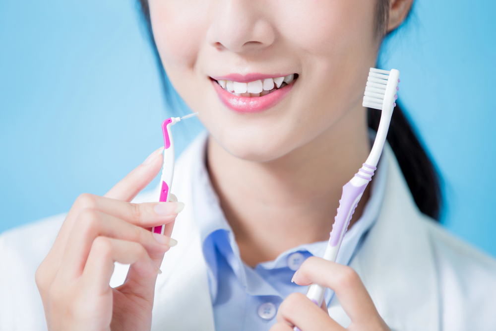 A woman in a white coat smiles whilst holding a toothbrush in one hand and an interdental brush in the other, against a light blue background.