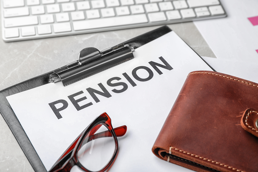 A clipboard holding a paper labelled PENSION sits on a desk with a pair of spectacles, a brown wallet, and part of a computer keyboard visible.