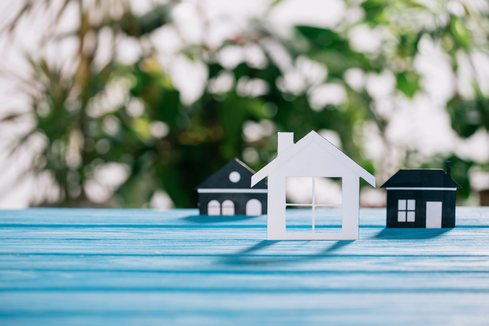 Three paper house models, one white and two black, sit on a blue wooden surface with a blurred green leafy background, representing the concept of home or property.