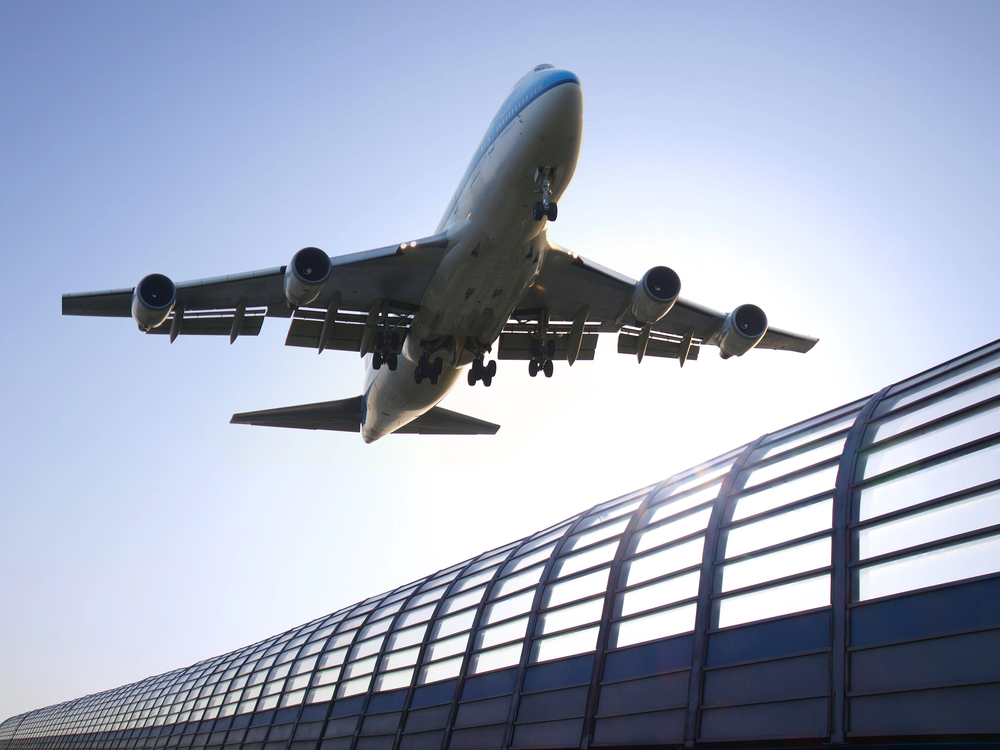 A large commercial aeroplane is flying low above a modern, curved, glass-covered structure, silhouetted against a clear blue sky.