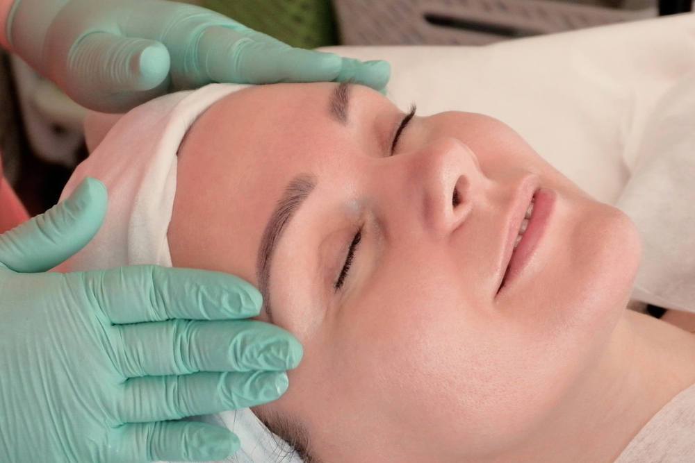 A person lies with eyes closed, smiling slightly, whilst someone wearing green gloves gently massages their forehead, suggesting a facial or spa treatment.