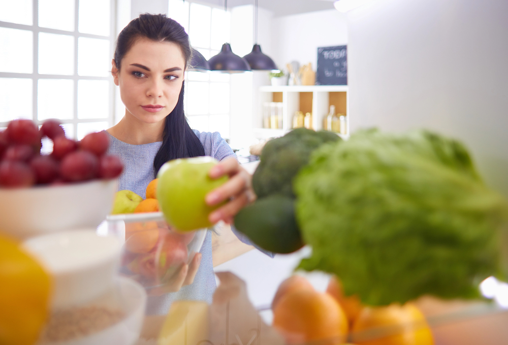 A woman looks into a fridge, reaching for a green apple among various fruits and vegetables, with a focused expression.
