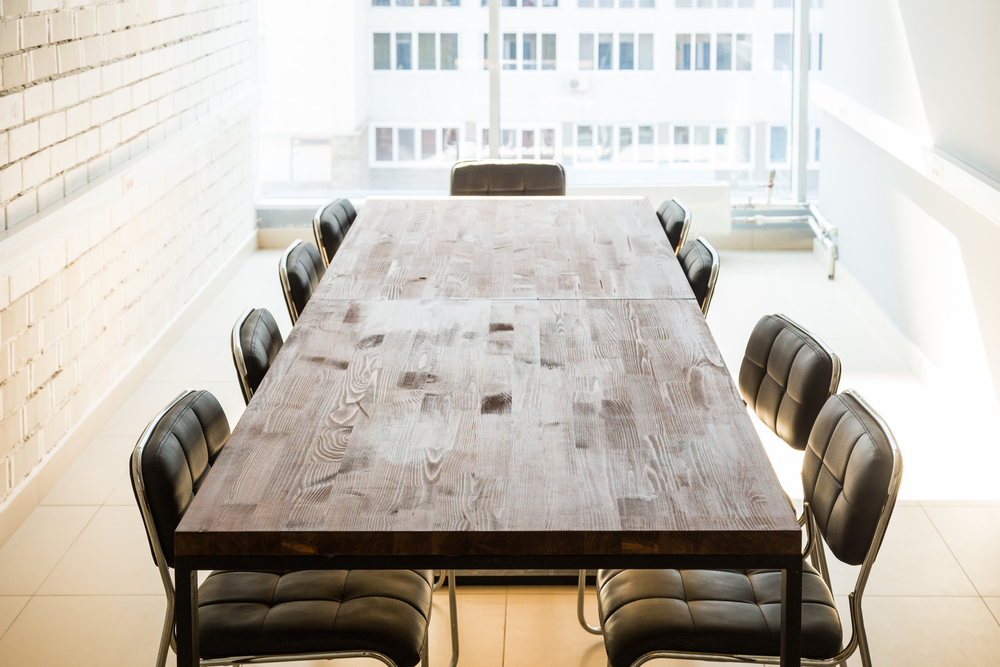 A sunlit conference room with a large wooden table surrounded by eight black padded chairs, set against white walls and large windows overlooking another building.