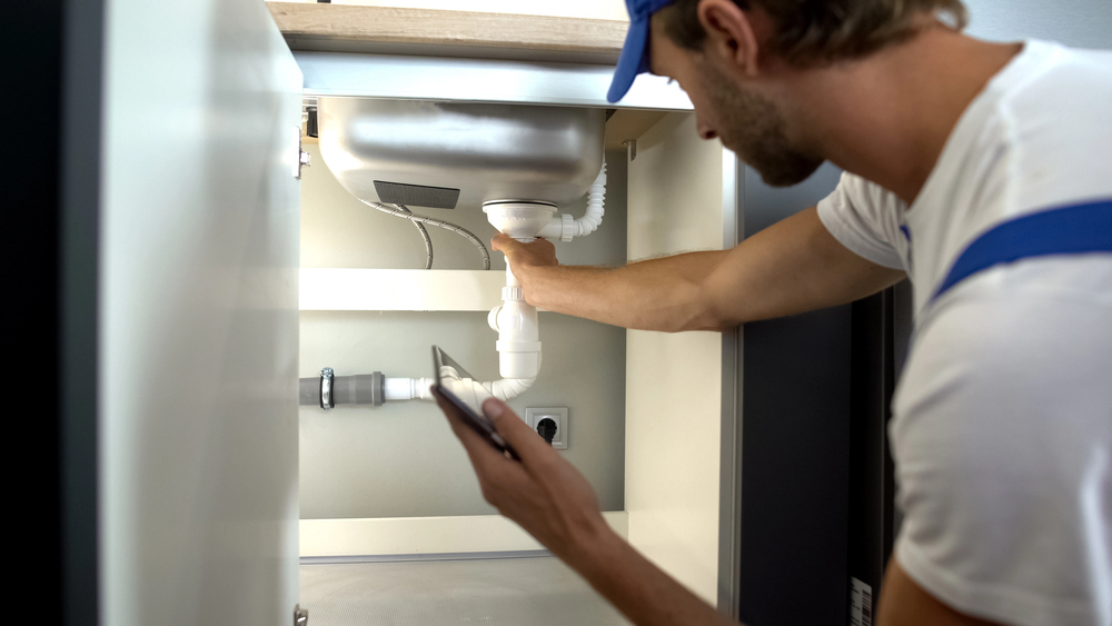 A plumber wearing a blue cap is fixing pipes under a kitchen sink, holding a mobile phone in one hand and adjusting the plumbing with the other hand.