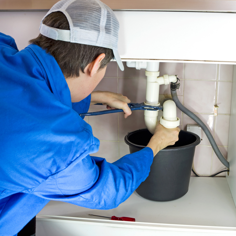 A plumber in a blue uniform uses a spanner to mend pipes under a sink, placing a black bucket below to catch water.
