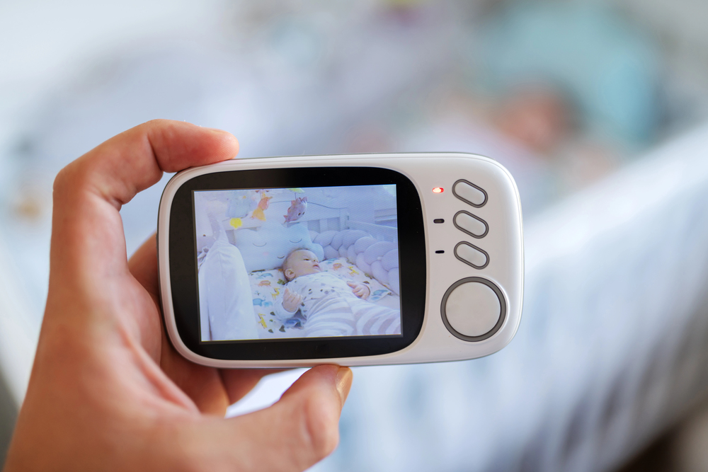A hand holds a baby monitor showing a live video of a baby lying in a cot, wearing striped clothes, with a blurred background of the actual cot and baby.