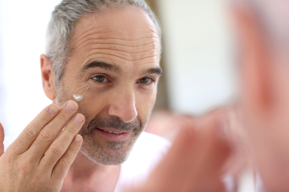 Middle-aged man with grey hair applies a small amount of cream to the skin under his eye whilst looking at his reflection in a mirror.