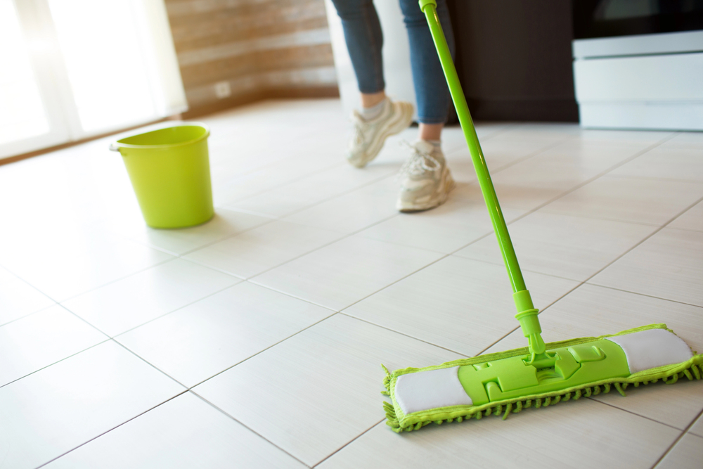 A person mops a tiled floor with a green mop, while a matching green bucket sits nearby. Only the person's legs and feet in trainers are visible.