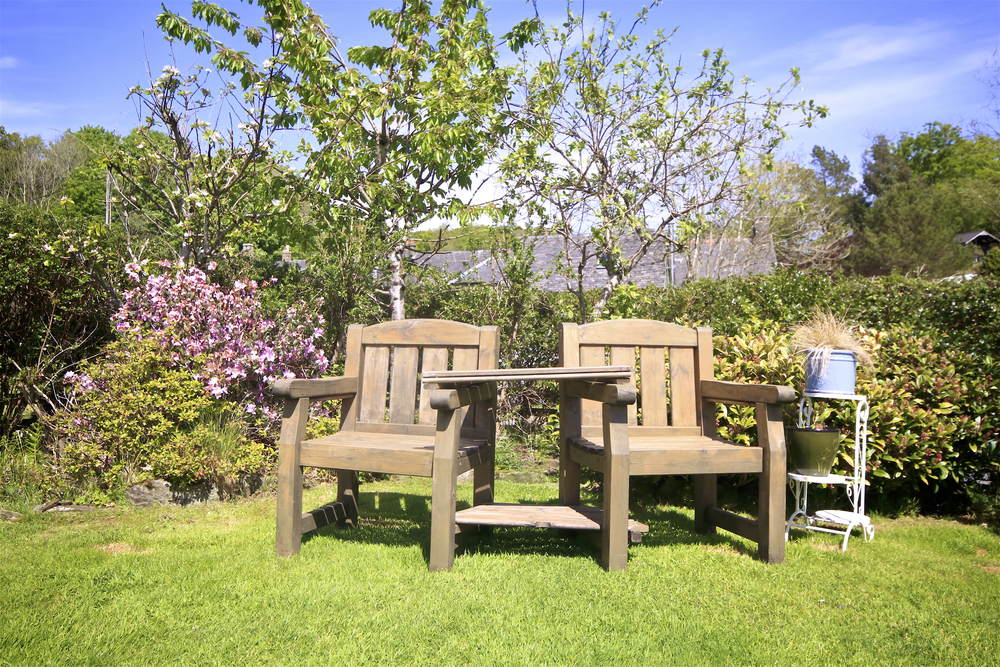 Two wooden garden chairs with a small table between them sit on green grass, surrounded by flowering plants, shrubs, and trees under a bright blue sky.