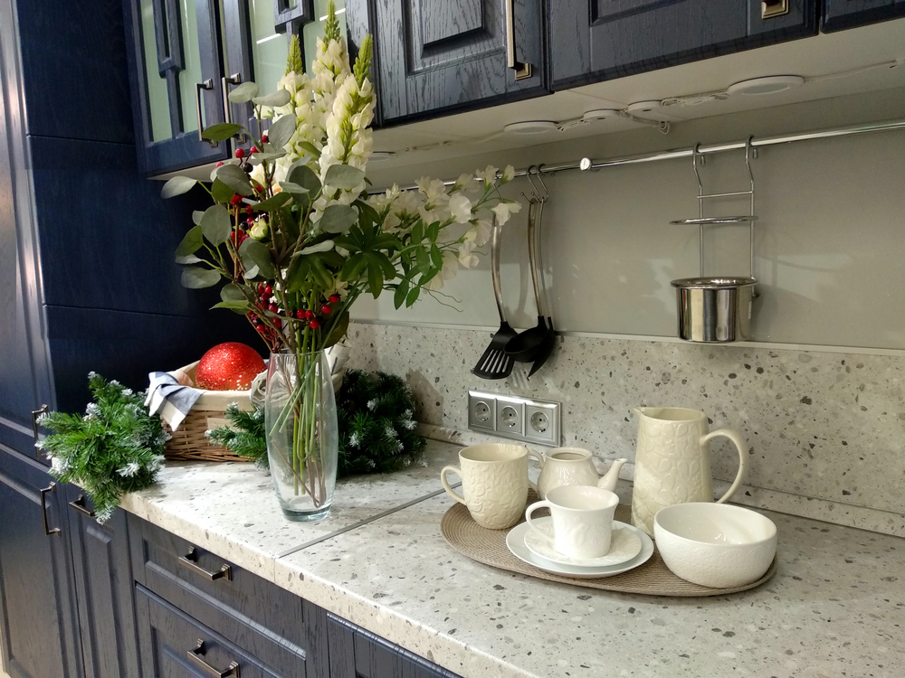 Modern kitchen worktop with a vase of white flowers, festive greenery, and a basket with a red bauble. White ceramic tea set on a tray sits on the granite worktop under dark cupboards.