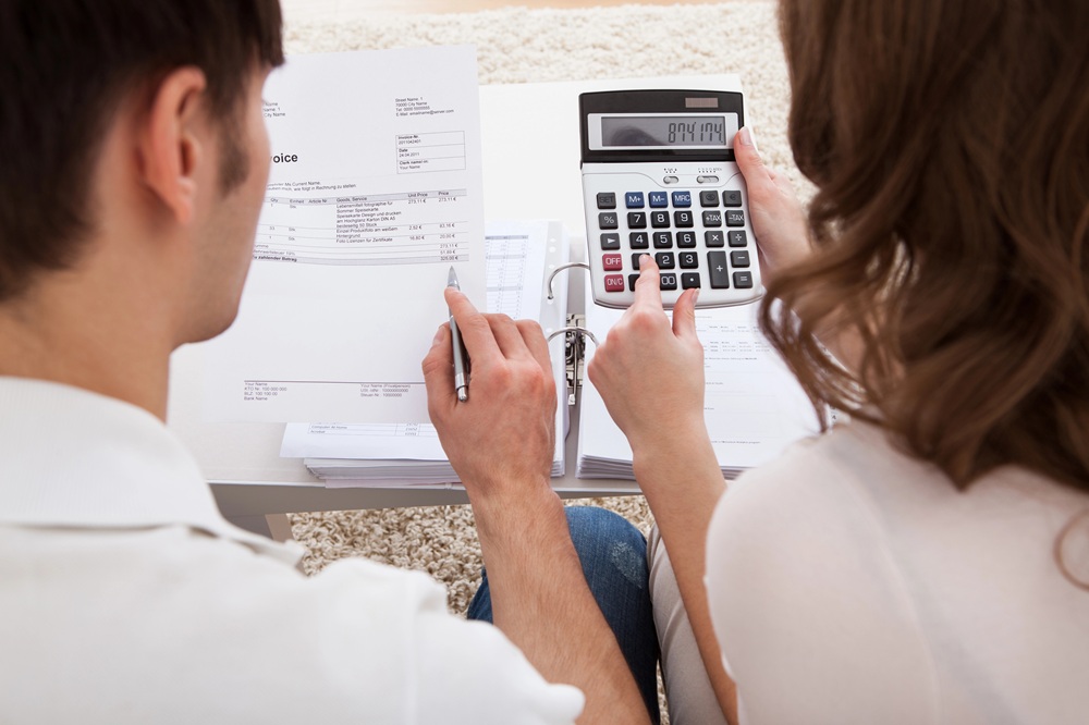 Two people sit at a table reviewing bills and papers; one holds a pen, the other uses a calculator, both focused on financial documents in front of them.