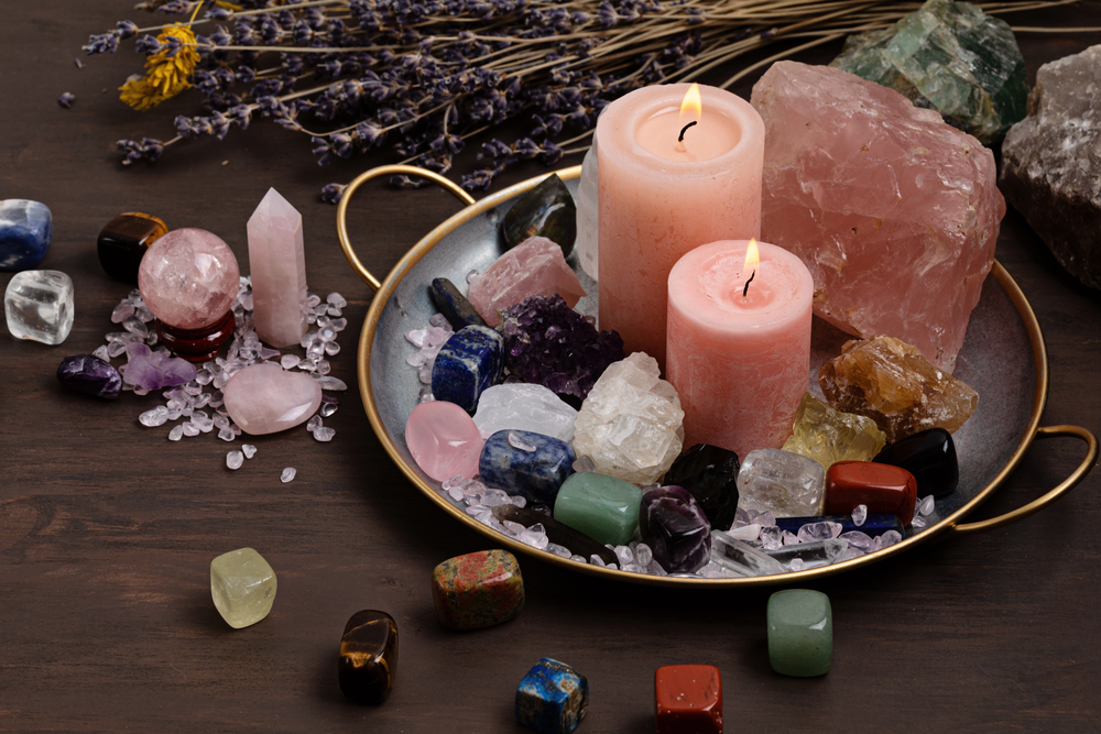 A round tray with two pink candles, surrounded by various colourful crystals and gemstones. Dried lavender is in the background on a dark wooden surface.