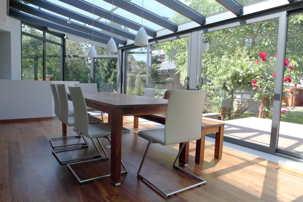 Modern dining area with a wooden table, white chairs, and large glass windows and ceiling overlooking a green garden with red flowers. Sunlight fills the room.