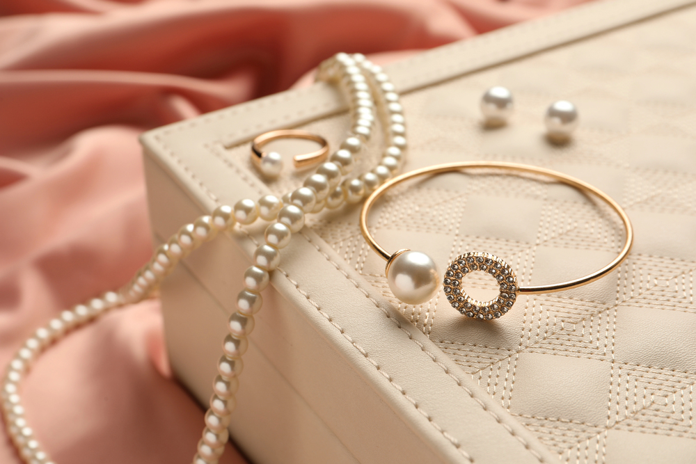 A gold bracelet, a pearl ring, and a pearl necklace rest on a quilted jewellery box, with loose pearls and pink fabric in the background.